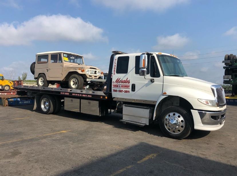 Tan Toyota Land Cruiser being transported on a white tow truck under a blue sky.