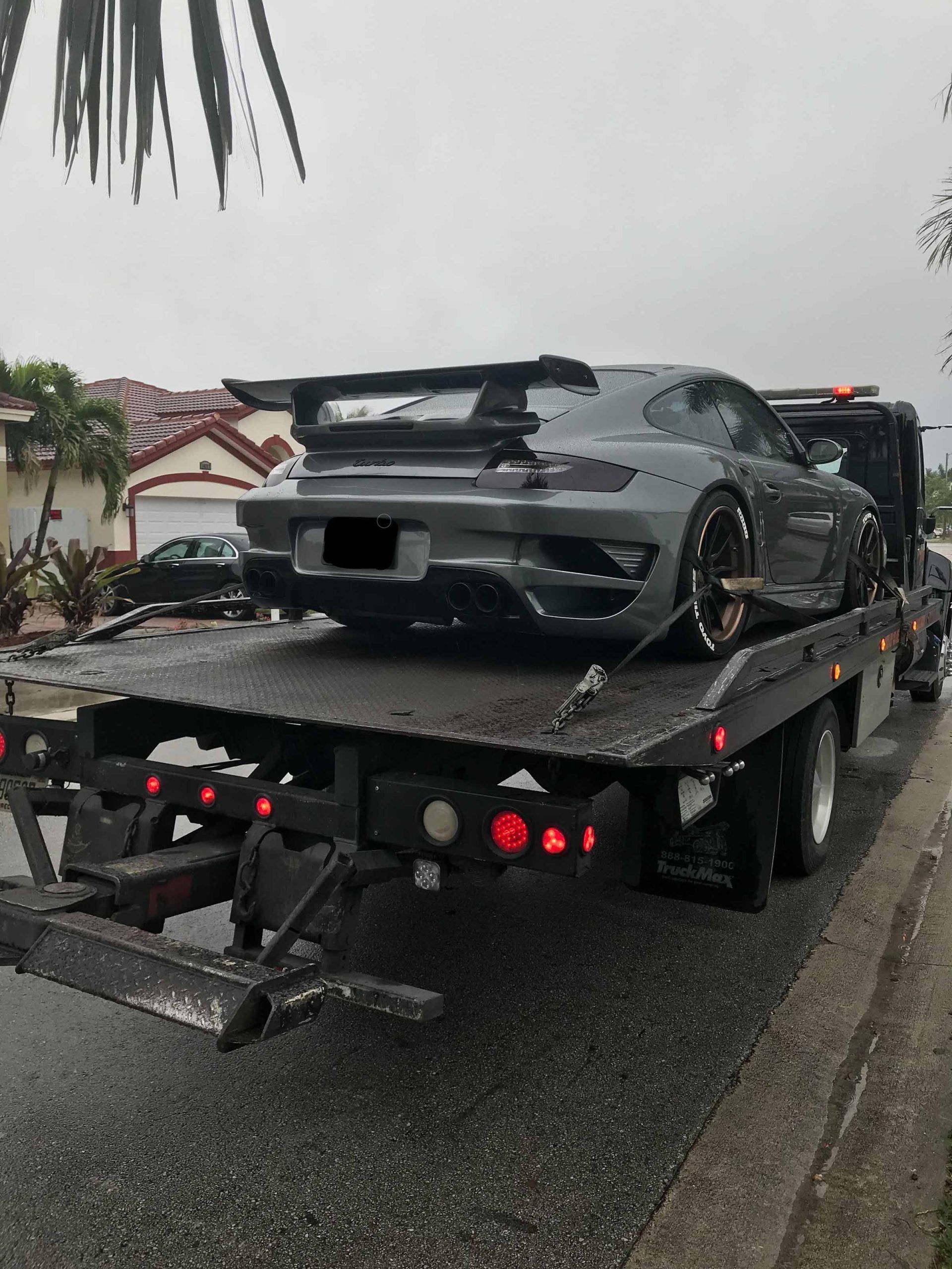 Gray sports car on a tow truck, likely broken down. Exterior daytime shot on a wet street.