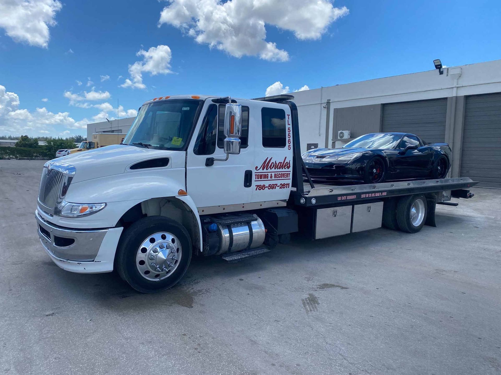 White tow truck with black sports car on the flatbed, parked outdoors under a blue sky.