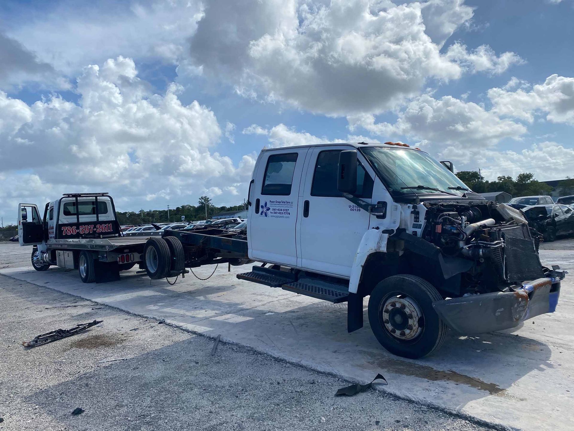 White truck chassis with cab missing front parts, parked on a concrete lot. Another truck in the background.