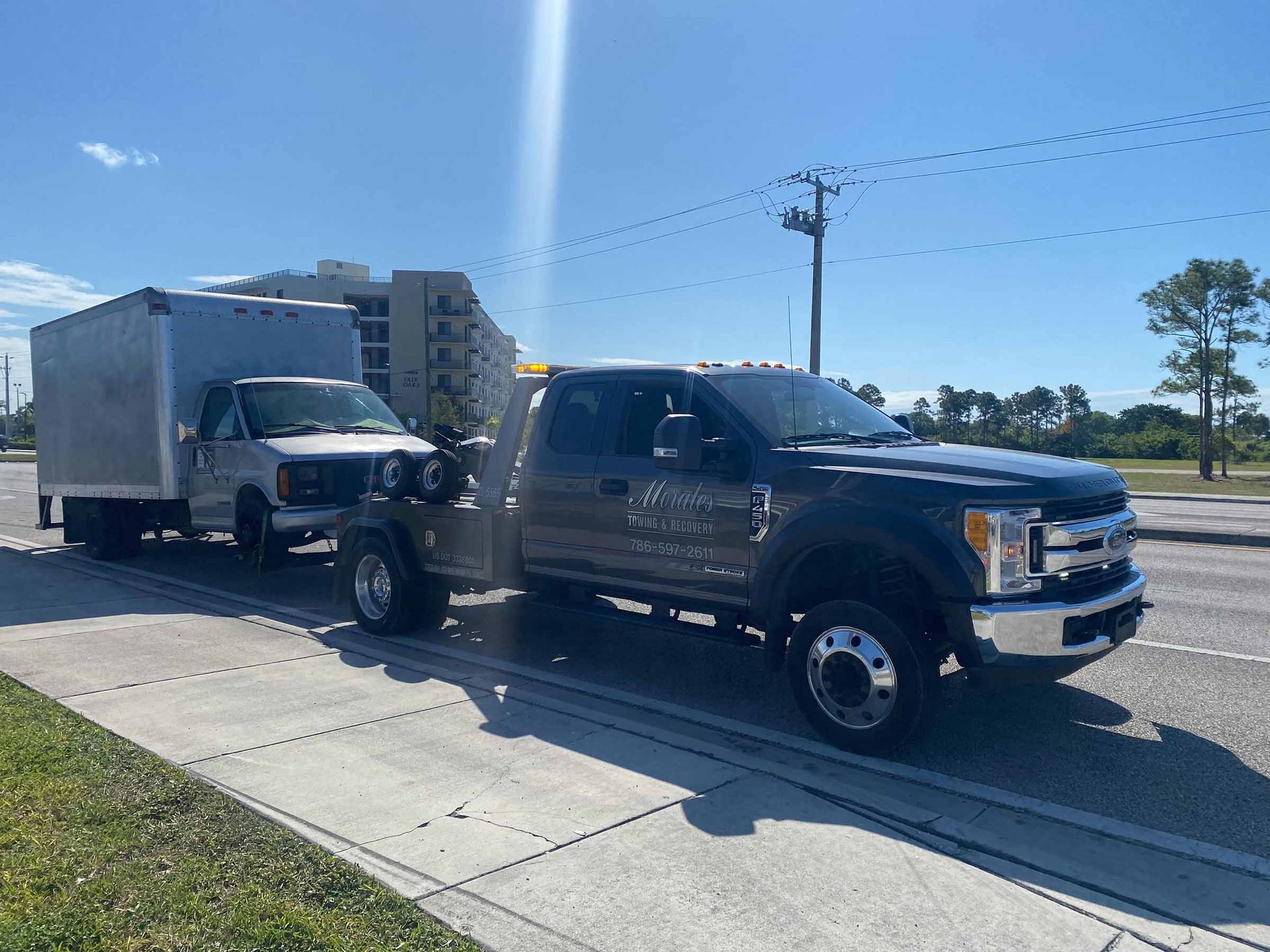 A dark gray pickup truck towing a box truck on a paved road under a sunny sky.