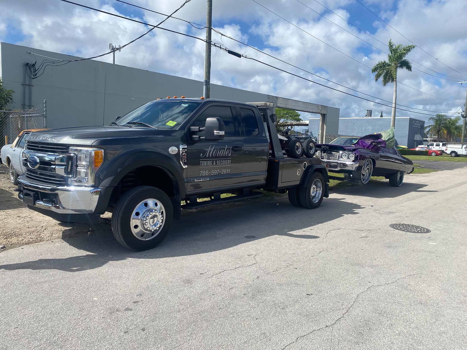 Dark grey tow truck with a trailer carrying a purple car parked on a street near industrial buildings.