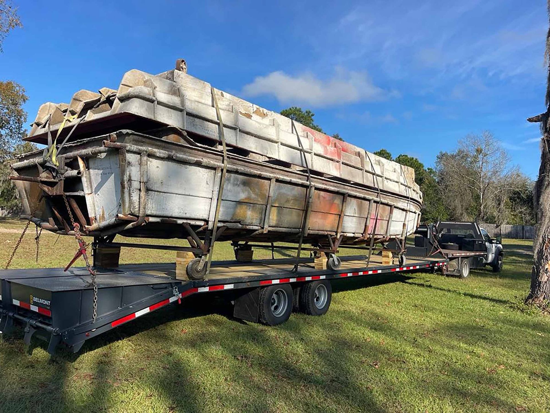 A large, metal pontoon boat loaded on a flatbed trailer, being pulled by a truck on grass.