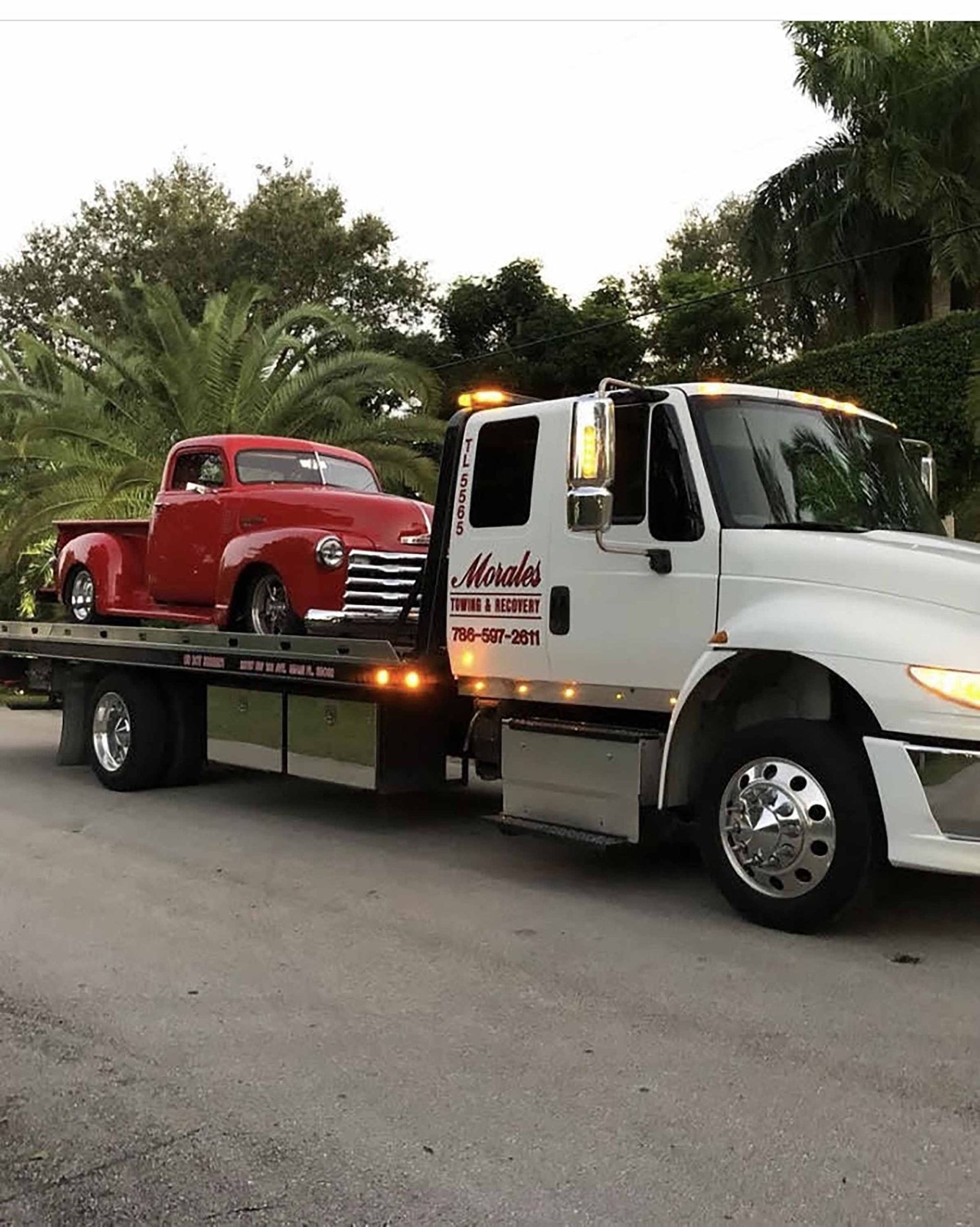 Red vintage pickup truck being towed by a white tow truck on a paved road.
