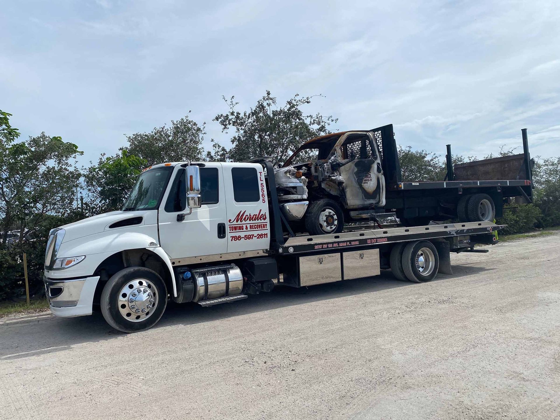 White tow truck hauling a burned-out vehicle on a gravel road, overcast sky in background.