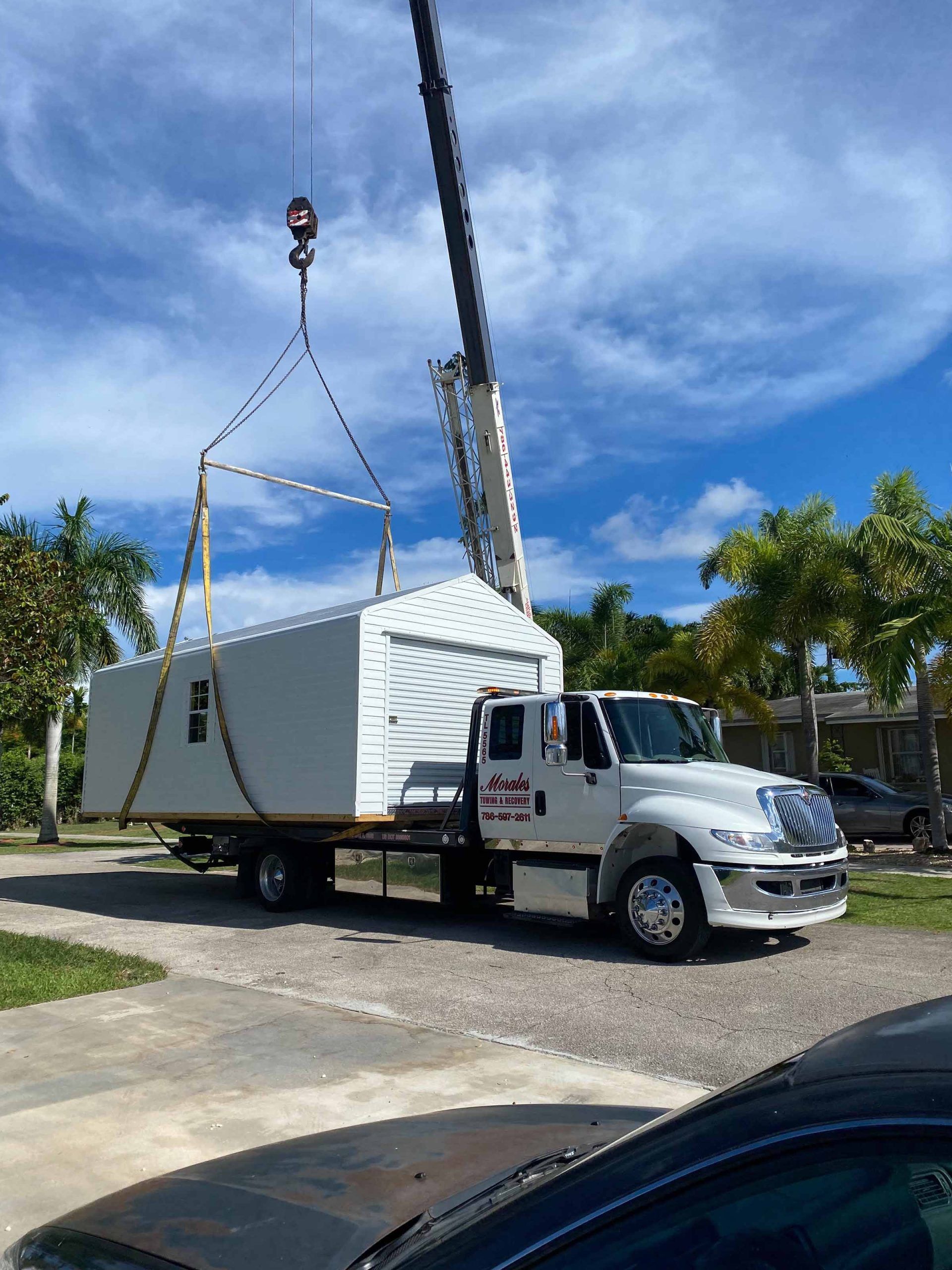 A crane lifting a white shed off a truck on a driveway under a blue sky.