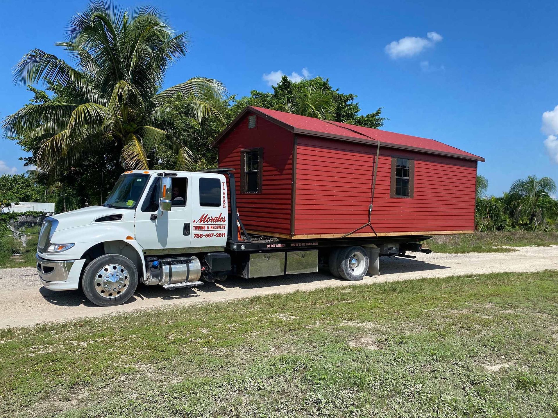 Red shed loaded on a white tow truck on a dirt road. Bright, sunny day.