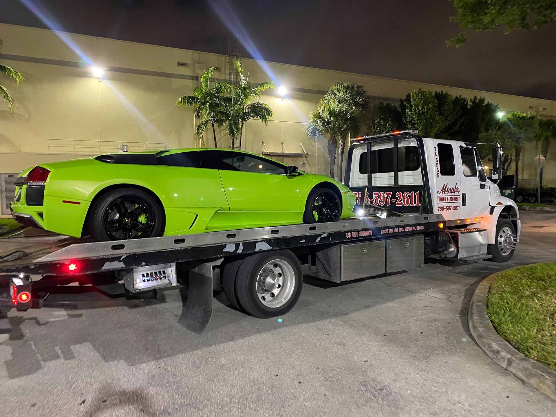 Green Lamborghini on a tow truck at night, parked near a building with lights.
