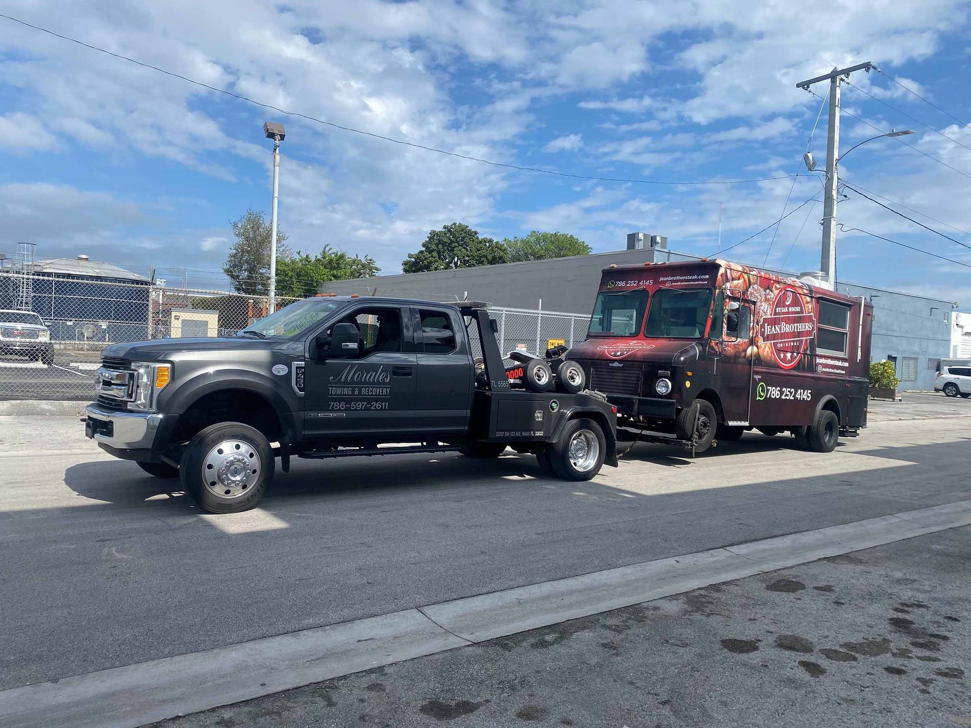 Gray tow truck towing a food truck on a street under a cloudy sky.