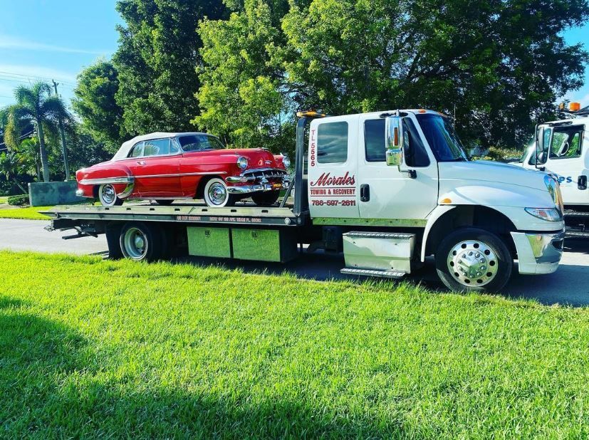 Red classic car on a flatbed tow truck. Truck is white with business logo; parked on grass near road.