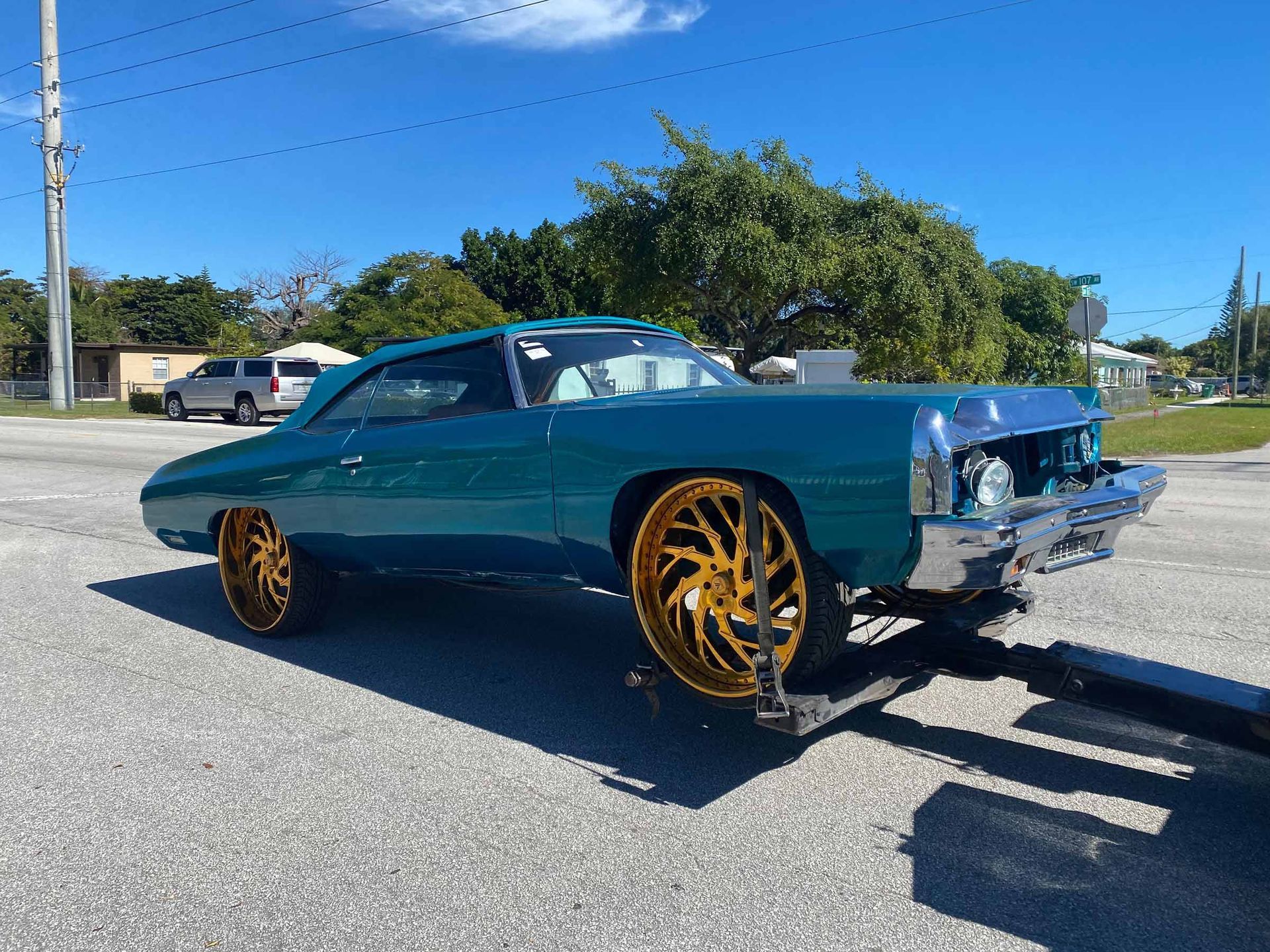 Teal classic car with gold rims on a flatbed tow truck outdoors on a sunny day.