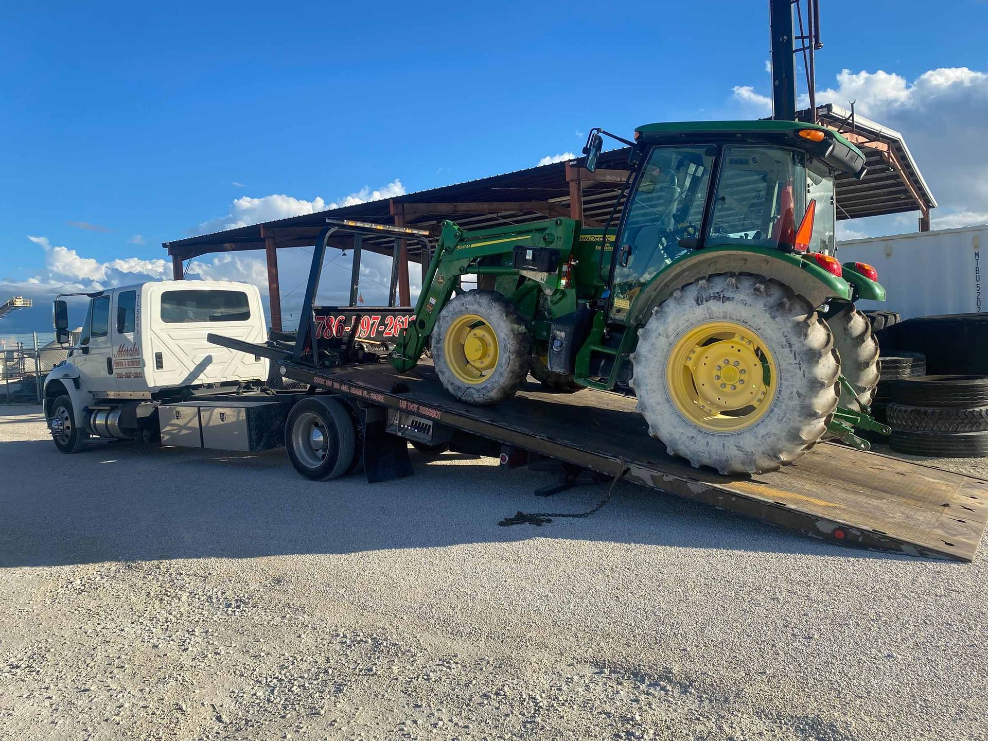 Tractor on a flatbed tow truck outdoors on a gravel lot under a partly cloudy sky.