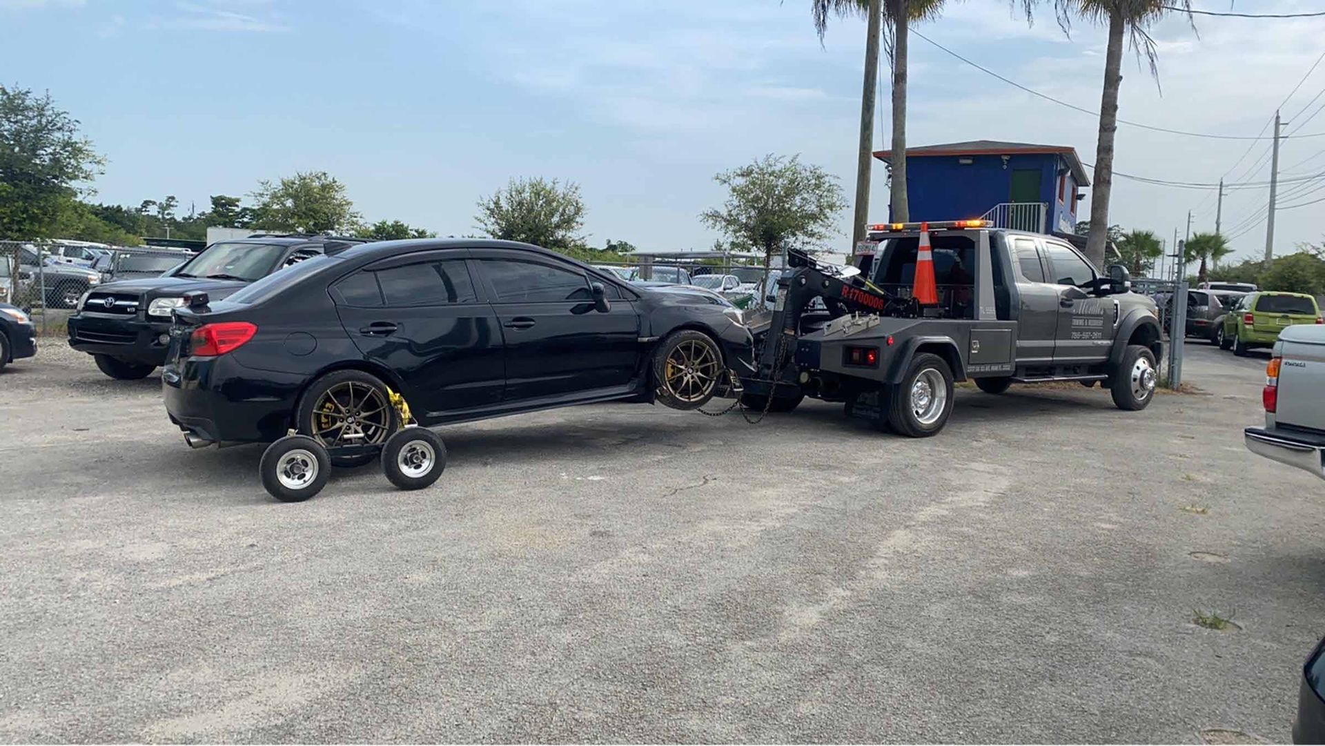 Black car being towed by a tow truck on gravel, wheels on dollies, outside on a sunny day.