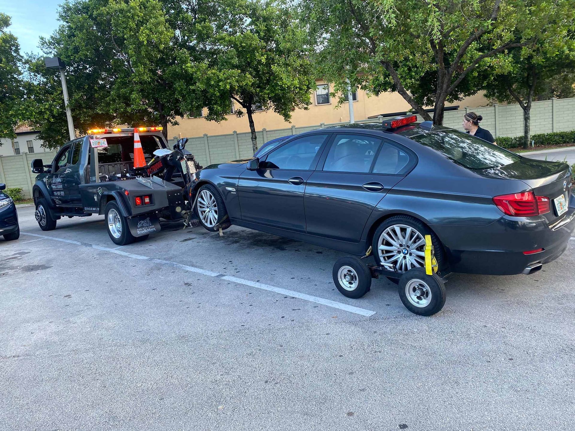 Tow truck towing a dark gray BMW sedan in a parking lot. Rear wheels are on dollies.