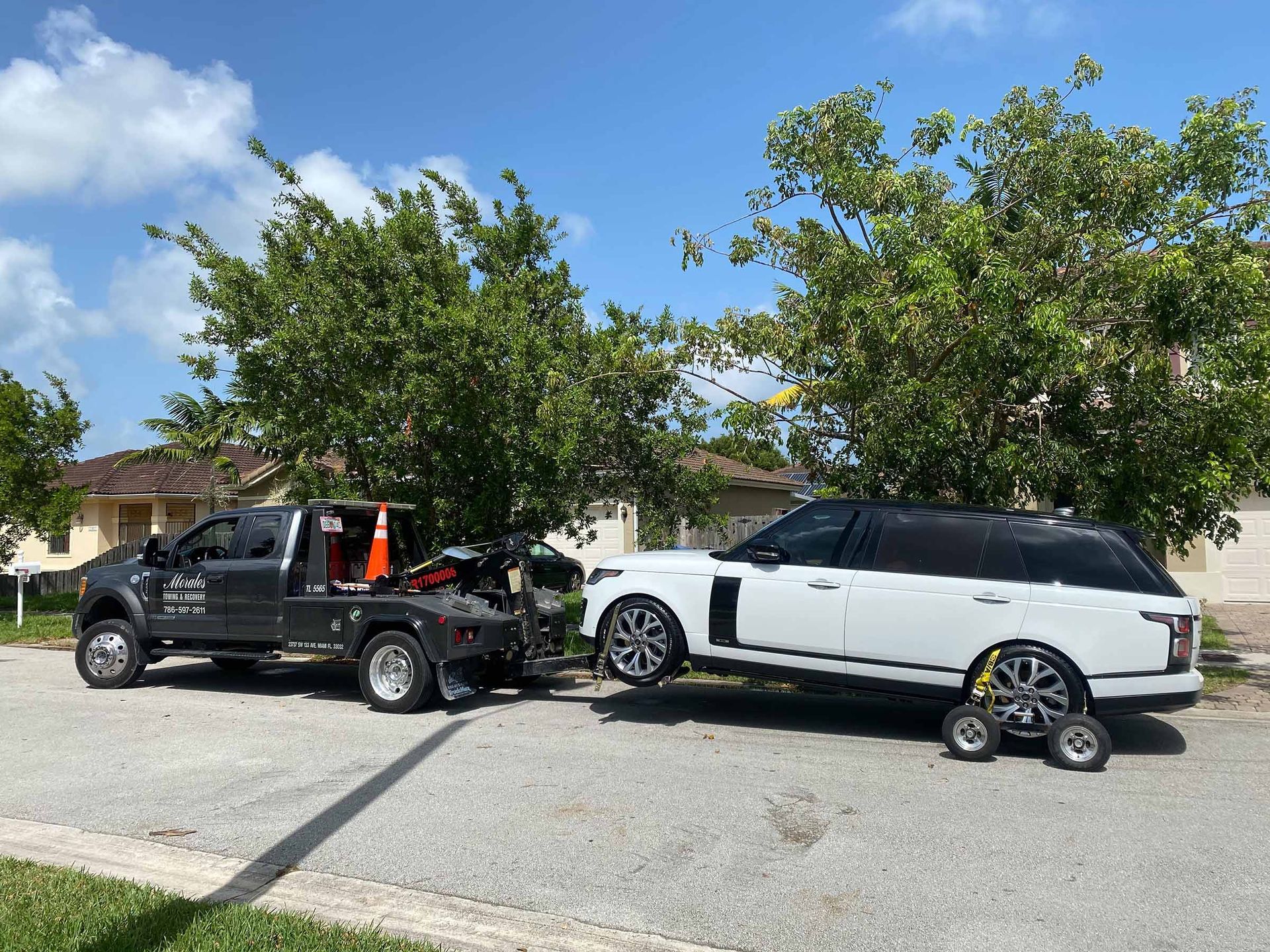A black tow truck towing a white Range Rover on a sunny street, next to houses and trees.