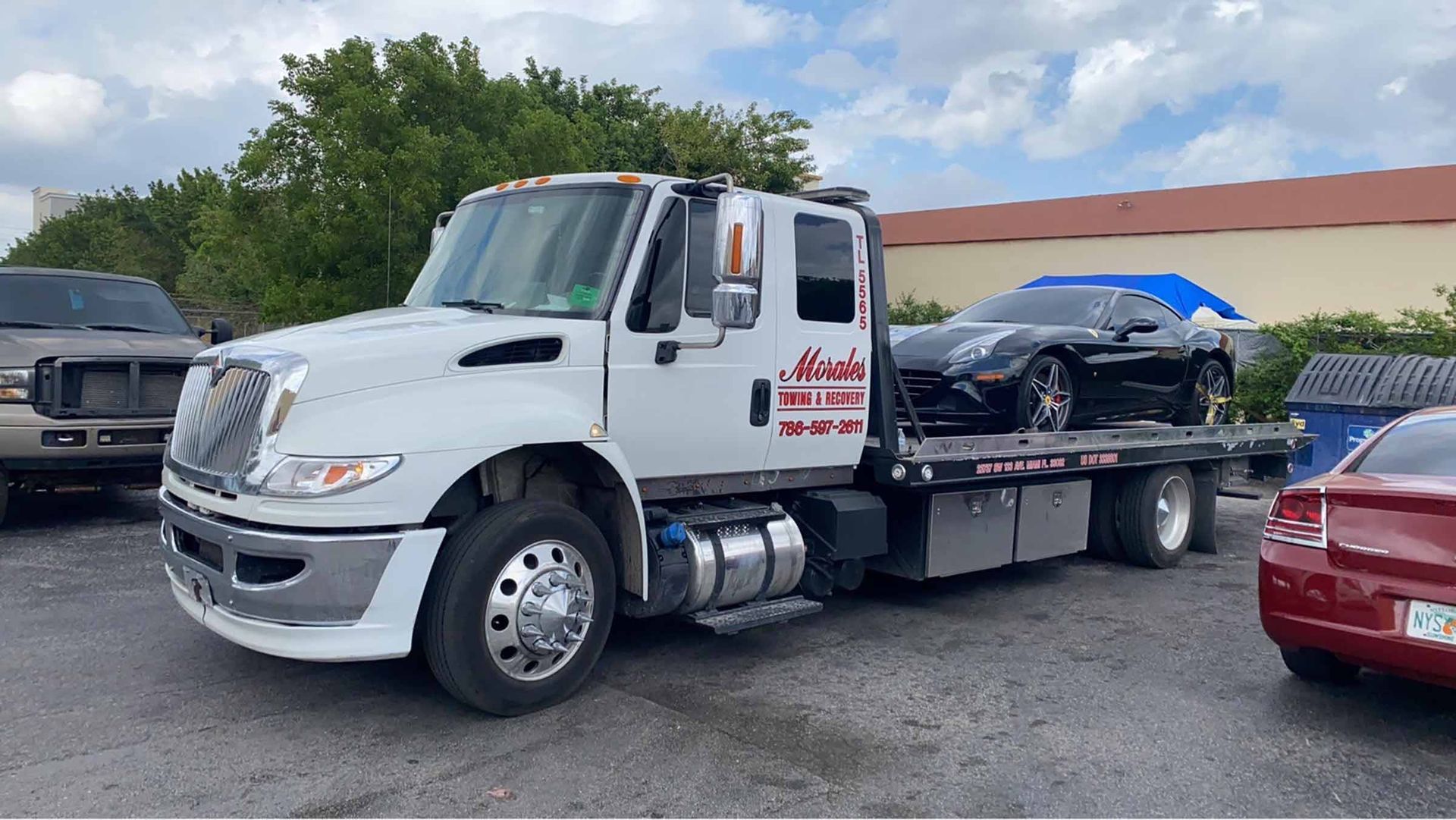 White tow truck carrying a black sports car on a flatbed. Outdoors, cloudy sky.