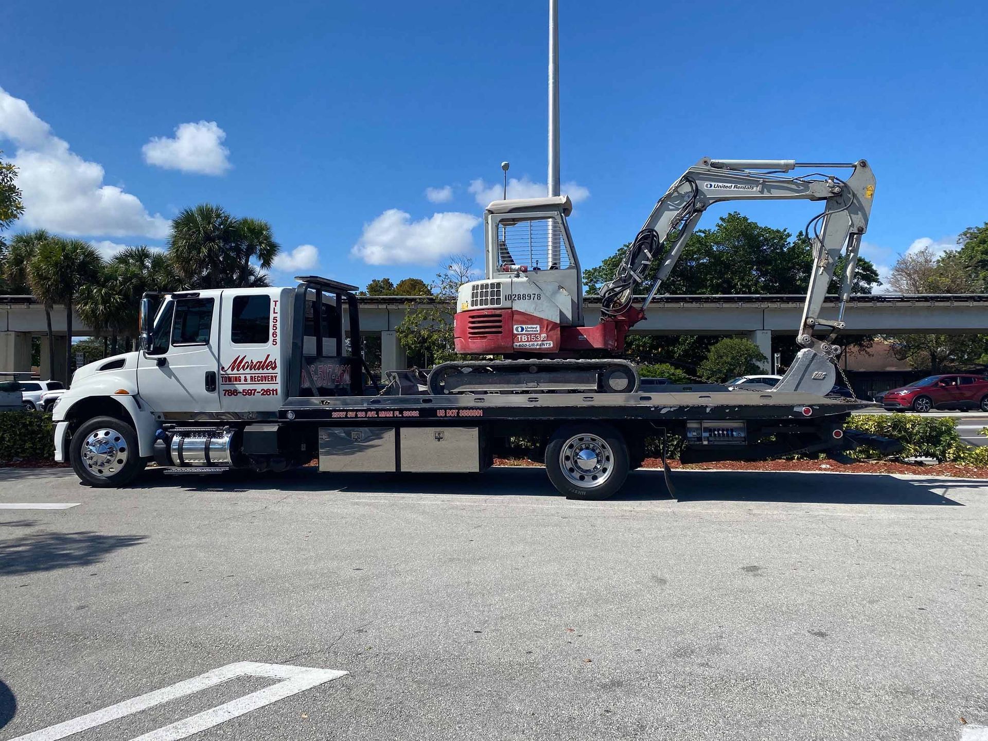White tow truck transporting a small red and white excavator on a sunny day.