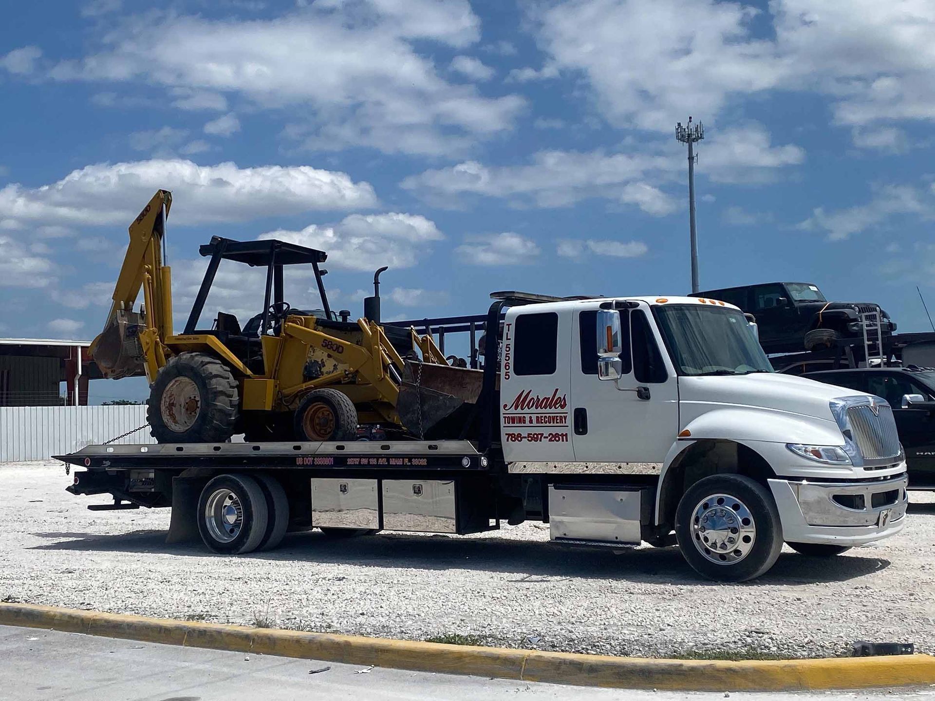 Yellow backhoe on white tow truck under cloudy sky.