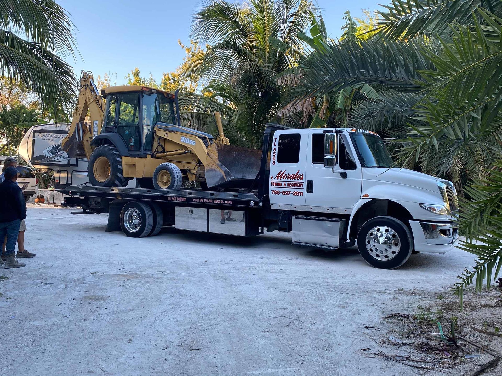 A white flatbed tow truck with a yellow backhoe loaded on it, in a sandy area.
