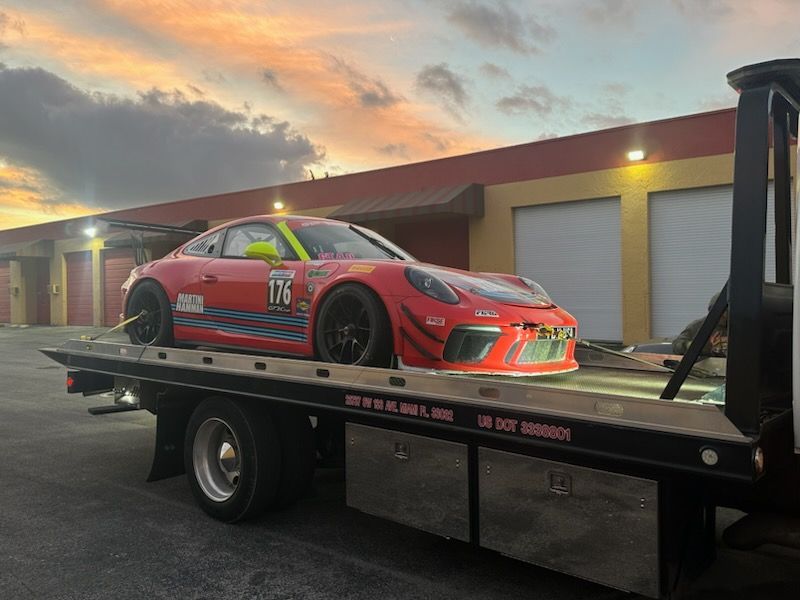 Red race car on a flatbed tow truck in front of a building at sunset.