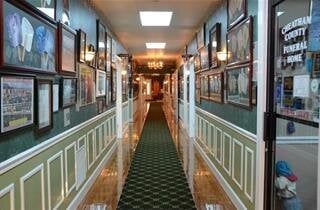 Long, narrow hallway in a funeral home. Green carpet, framed art, and decorative wall panels line the walls.