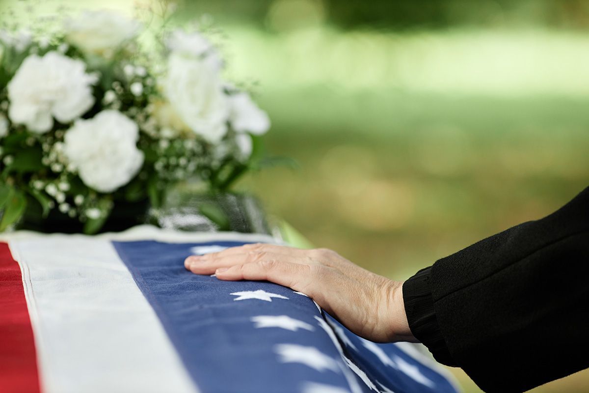 Hand resting on a casket draped with the American flag, white flowers in background.