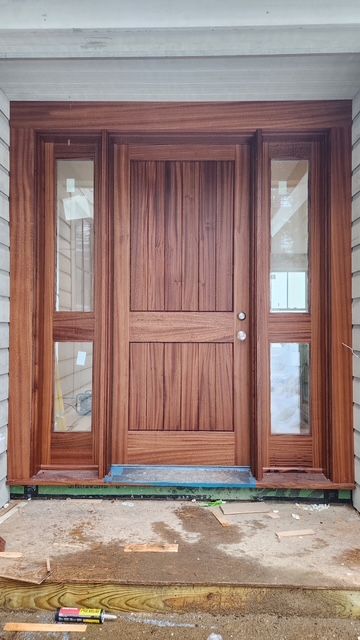 Wooden front door with sidelights, freshly installed, under a covered porch.