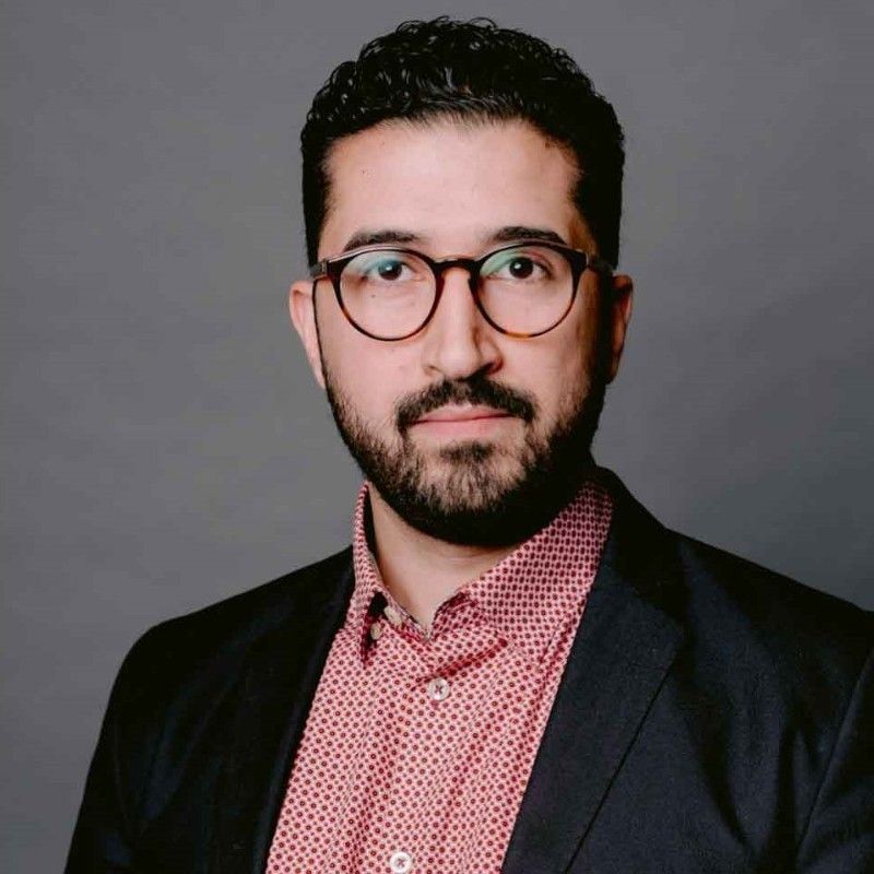Un homme avec une barbe et des lunettes porte un costume et une chemise rouge.