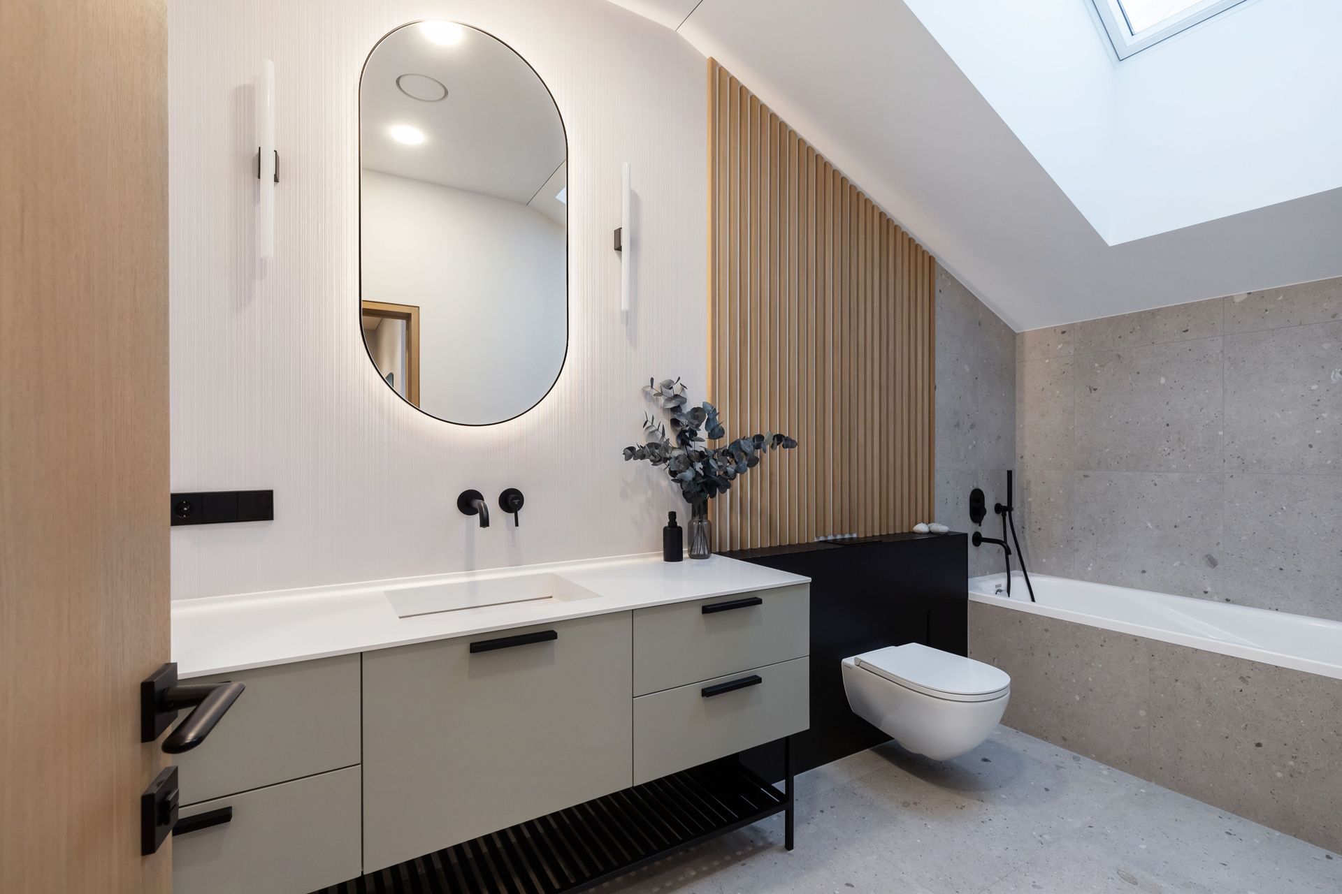 Modern bathroom with oval mirror, grey vanity, toilet, and bathtub under skylight.