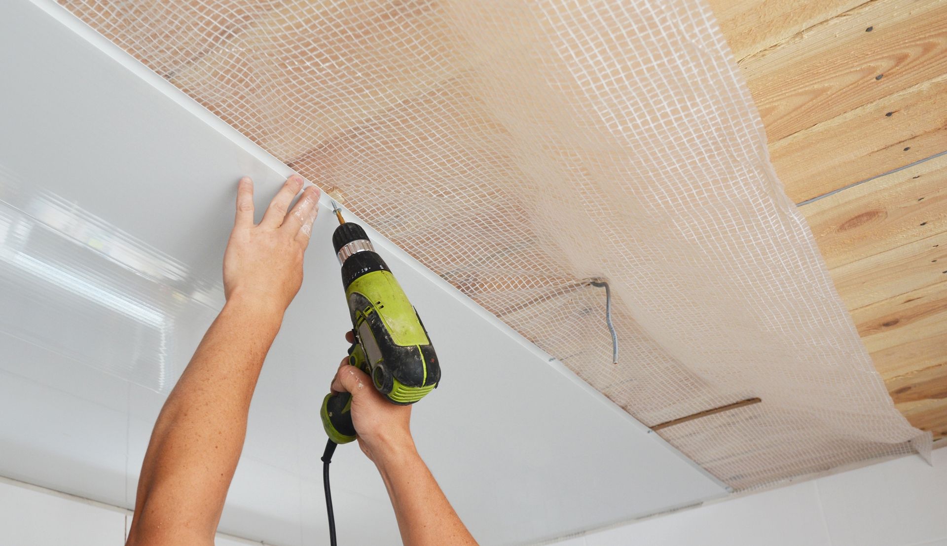 Person using a drill to install a white ceiling panel, brown netting visible above.