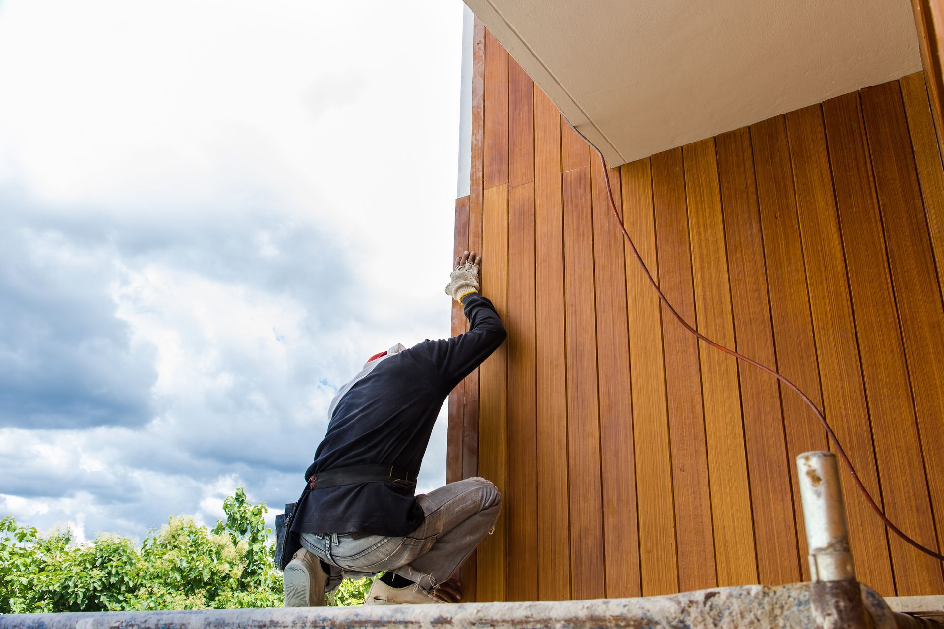 Construction worker installs vertical wooden siding on a building exterior, cloudy sky.