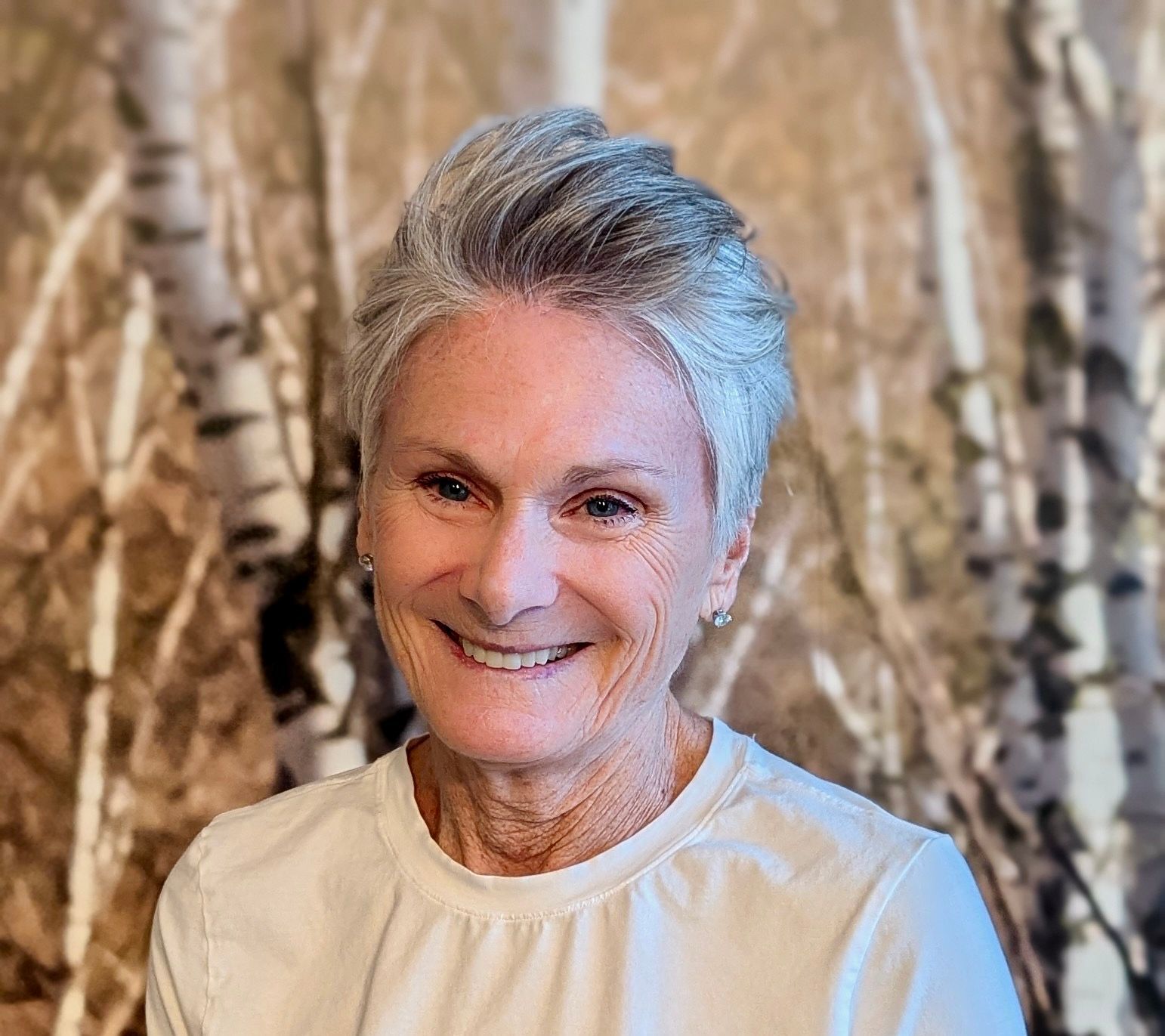 A woman in a white shirt is smiling in front of a painting of birch trees.