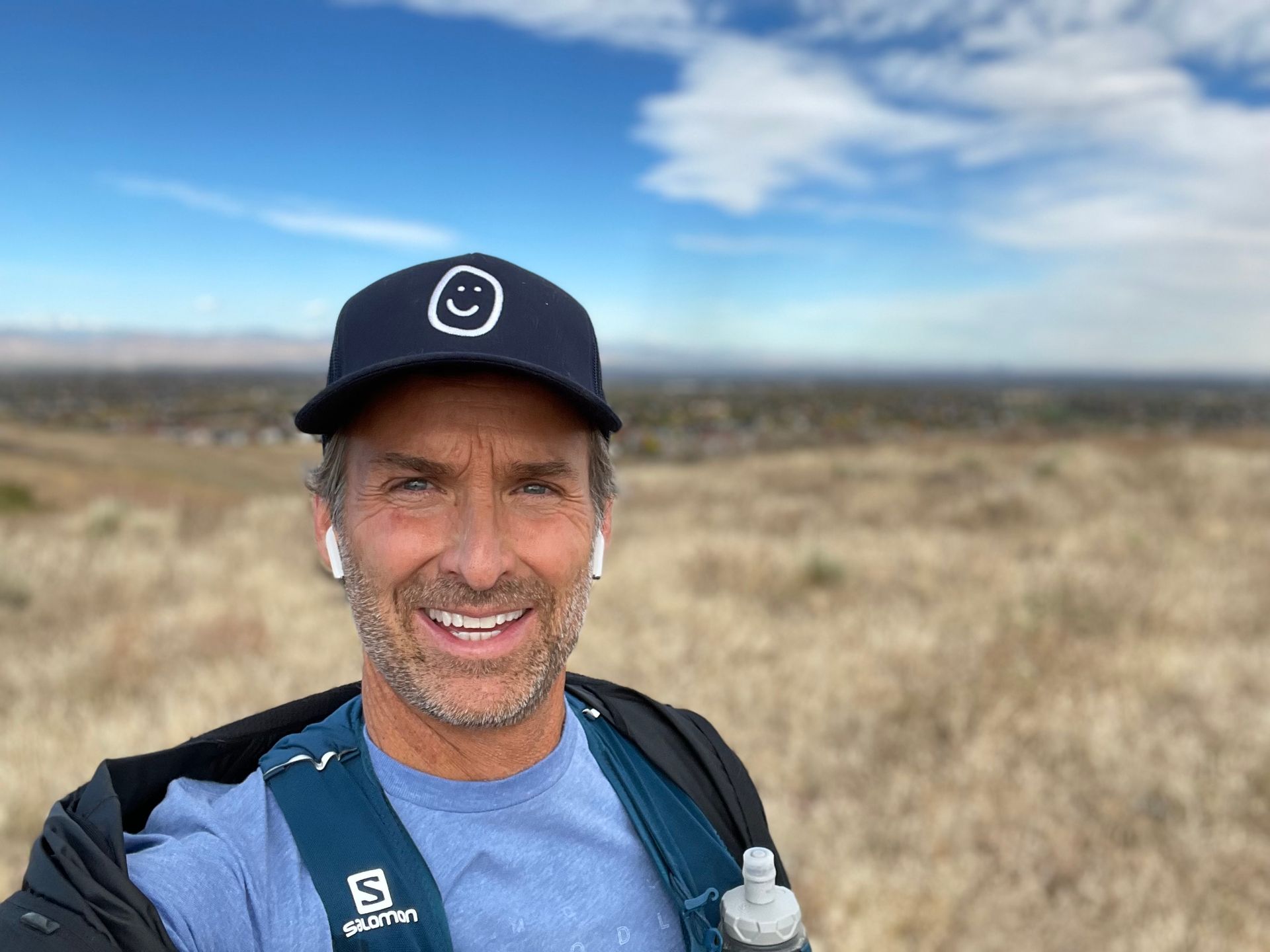 A man wearing a hat and a backpack is taking a selfie on top of a hill.