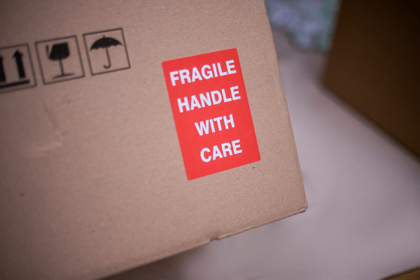 Bottom corner of a brown cardboard box with a red label on it, with white text stating 'FRAGILE HANDLE WITH CARE'.