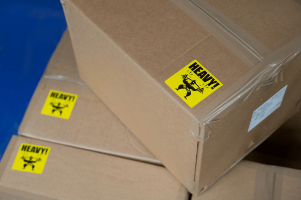 A number of cardboard boxes stacked on a pallet with a bright yellow label with the word 'HEAVY' in the corner of each one.