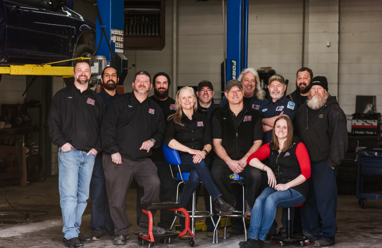 Group of people in black shirts posing in a workshop beside a seated woman in red and black. | Mears Automotive