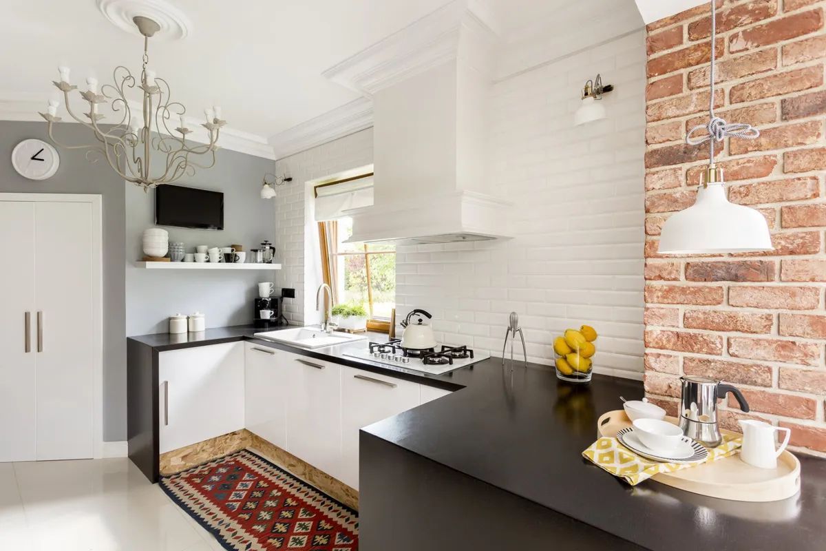 A kitchen with white cabinets , a black counter top , a brick wall and a chandelier.