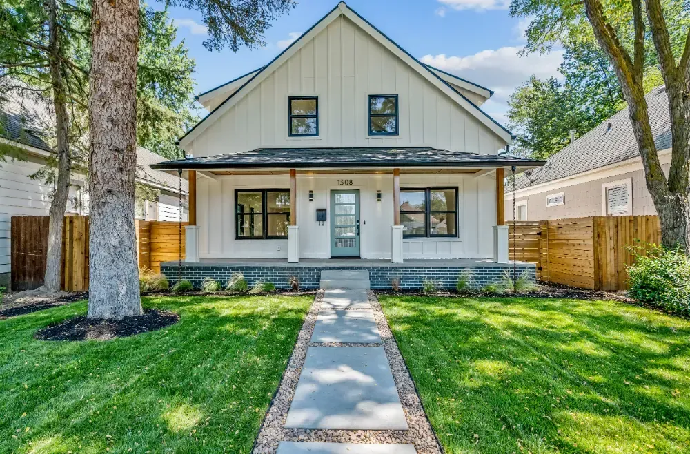 White farmhouse with black trim, blue-green door, porch, and walkway, set amongst green lawn and trees.