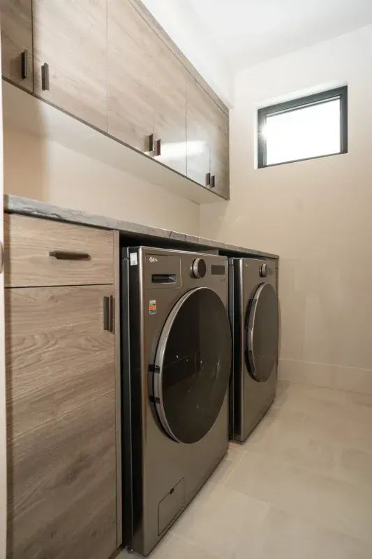 Laundry room with modern cabinets and silver washer and dryer units. A small window is on the right.