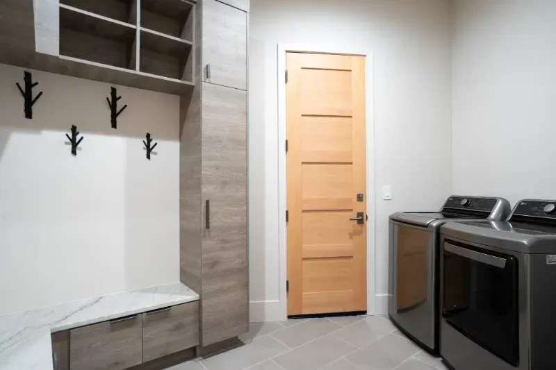 Laundry room with gray cabinets, washer/dryer, light wood door, and coat hooks.