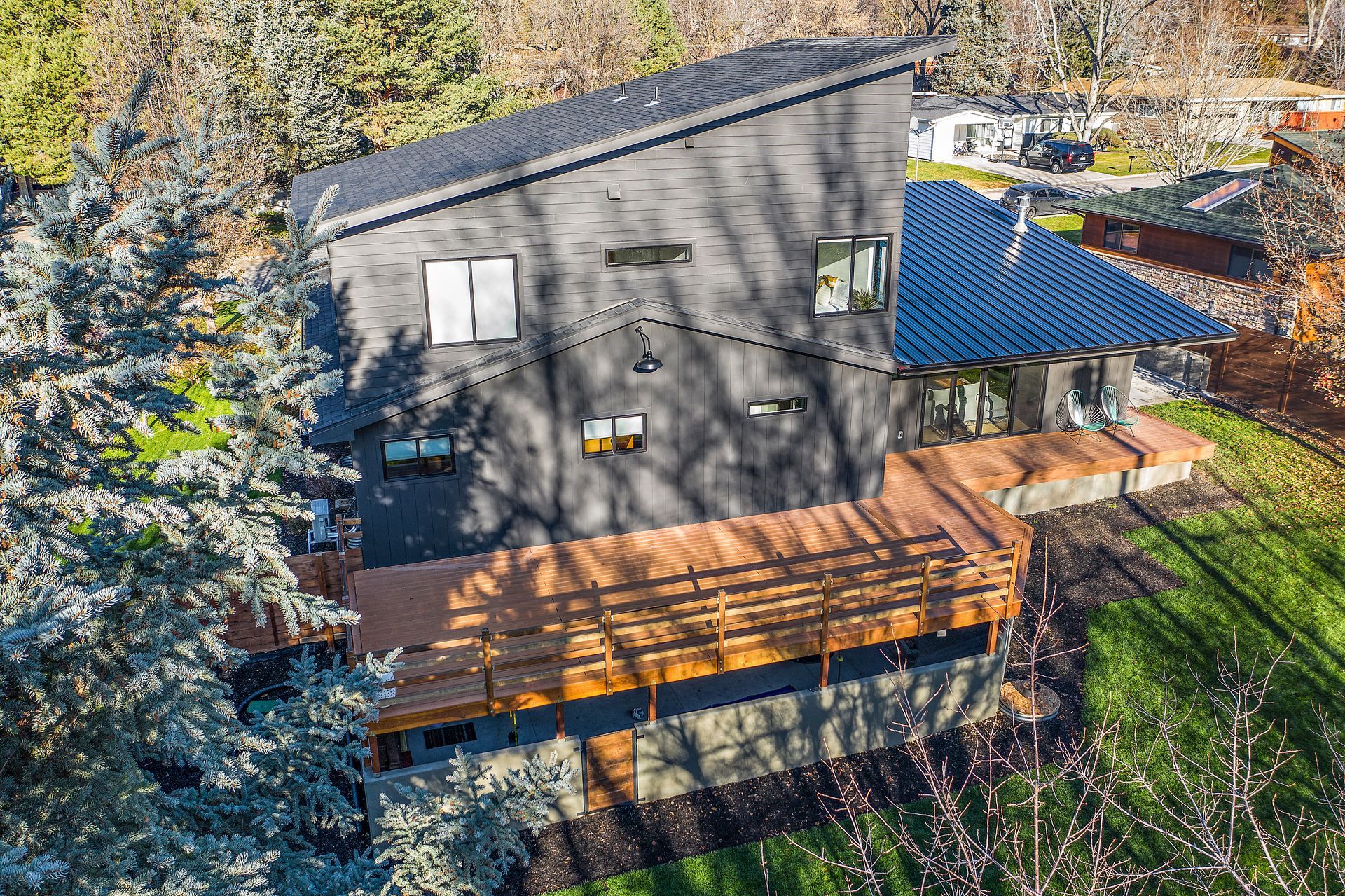 Modern two-story house with black exterior, angled roof, and wraparound wooden deck, surrounded by trees and grass.