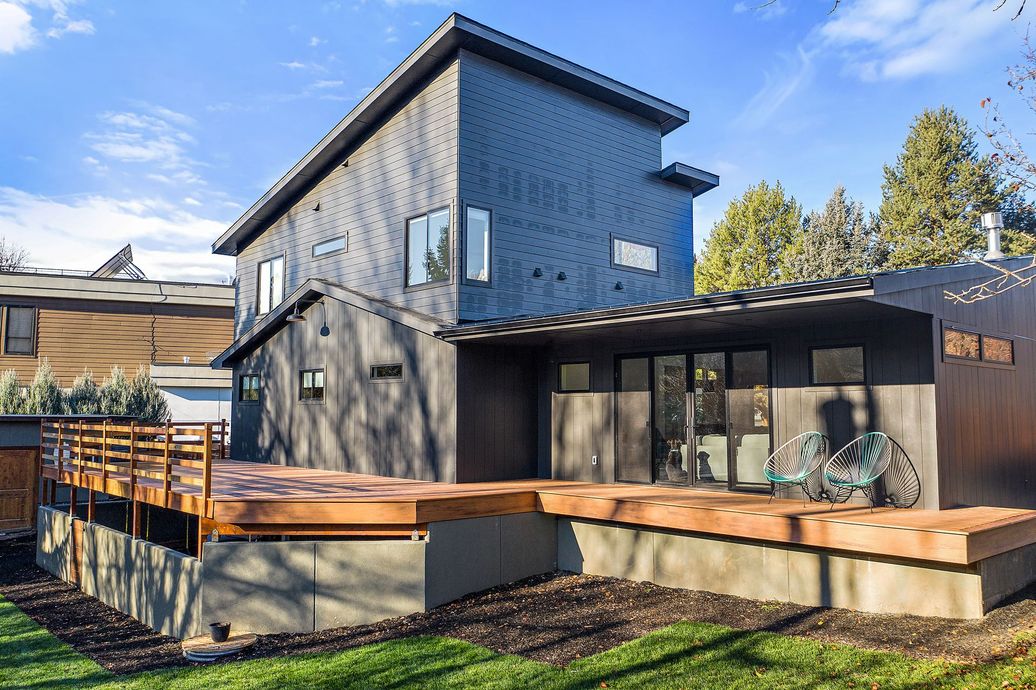 Modern two-story house with dark gray exterior, wooden deck, large windows, and a bicycle parked outside.