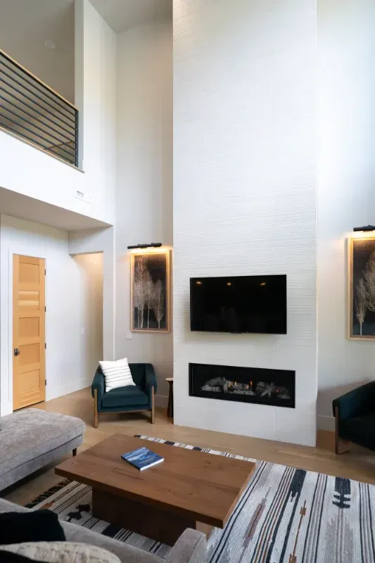 Living room with high ceiling, fireplace, TV, armchairs, artwork, and wooden coffee table.
