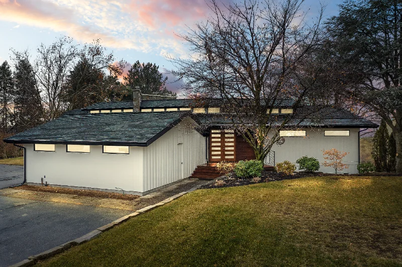 Mid-century modern home with a dark roof and white exterior. Grassy lawn, trees, and cloudy sky.