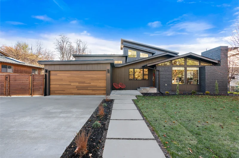 Modern brown house with wood garage door, concrete path, and blue sky.