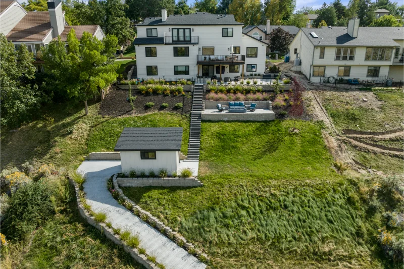 Backyard view of a white, multi-level house on a hillside with a shed and driveway.
