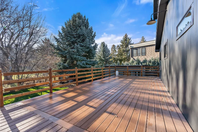 Wooden deck with brown planks and railing. Trees and blue sky visible. Building with dark siding on the right.