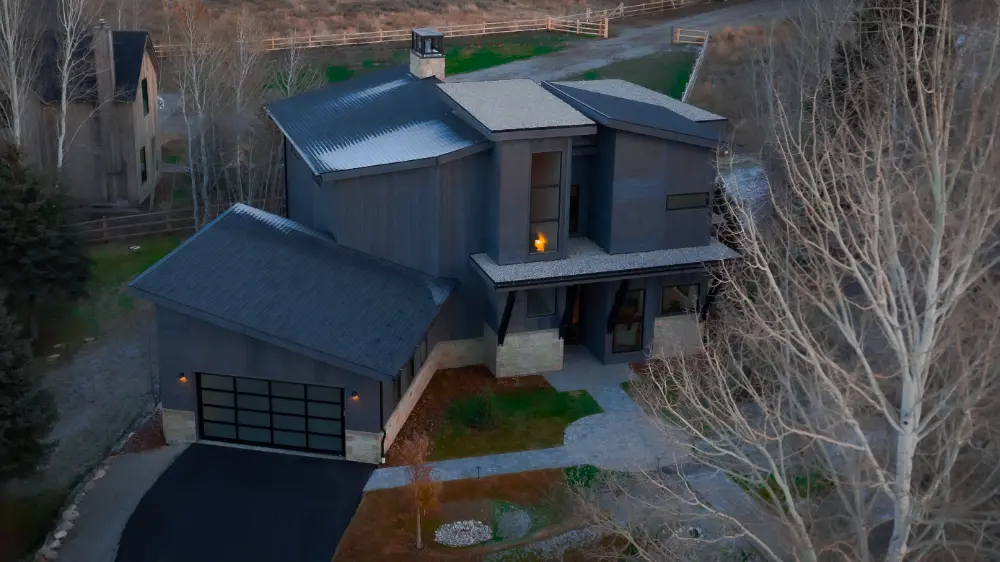 Modern gray house with dark roof and attached garage, on a tree-lined property.