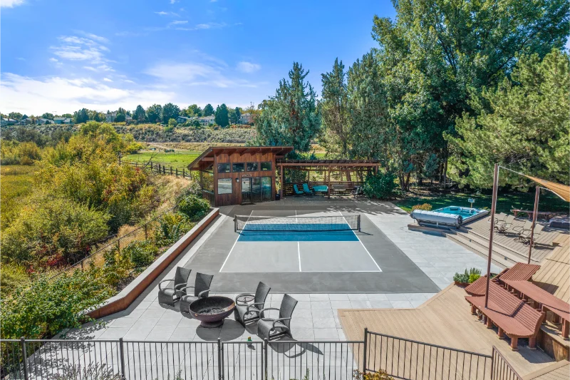 Backyard with pool, hot tub, deck, and shelter, surrounded by trees and a hill under a blue sky.