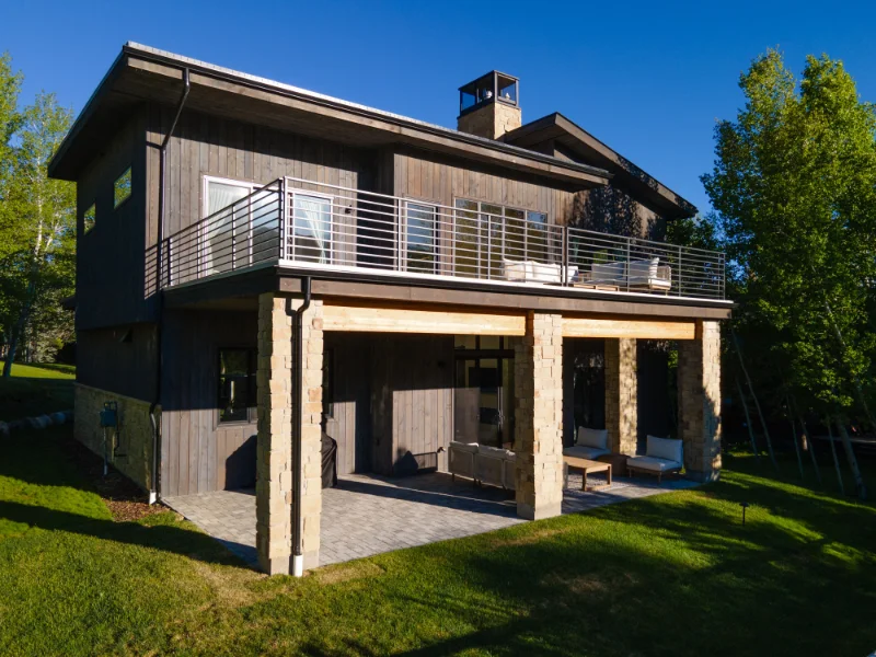 Two-story modern home with a covered patio and upper-level balcony; brown siding, stone columns, green lawn.